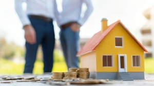 Two homeowners in blurry background with small yellow house and pile of coins before them contemplating DIY home project.