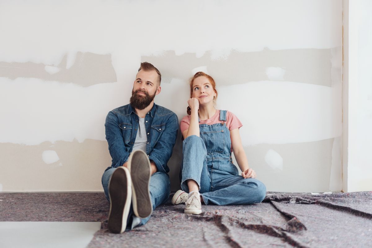 Man and woman seated on unfinished floor with bare wall behind them contemplating a DIY home project.
