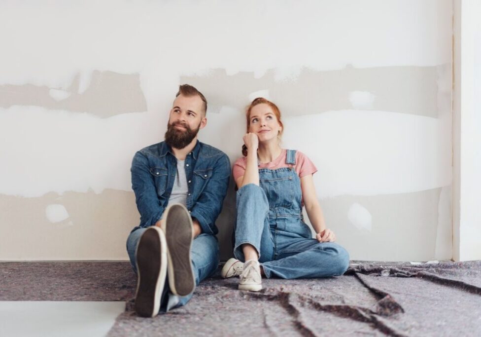 Man and woman seated on unfinished floor with bare wall behind them contemplating a DIY home project.